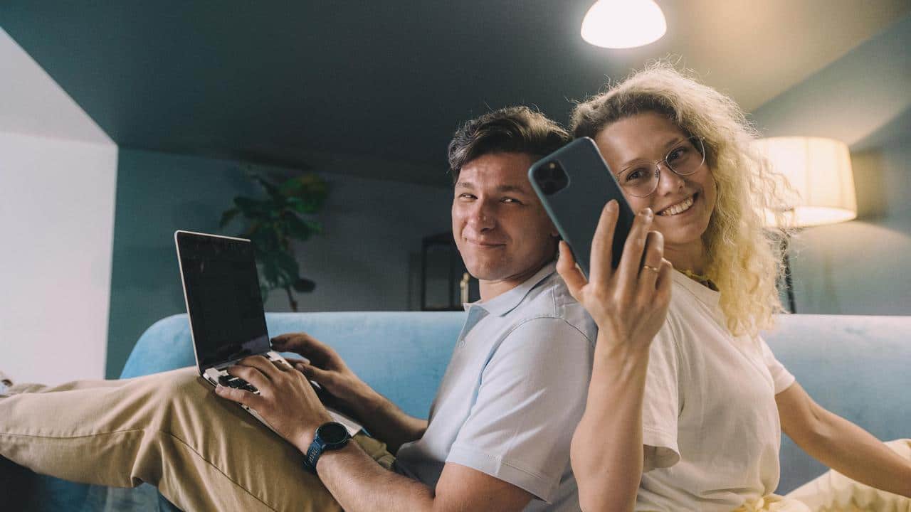 A couple sitting back-to-back on a couch using a laptop and smartphone.