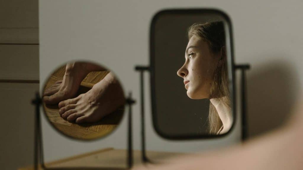A woman’s reflection in a mirror with her feet visible, appearing thoughtful.
