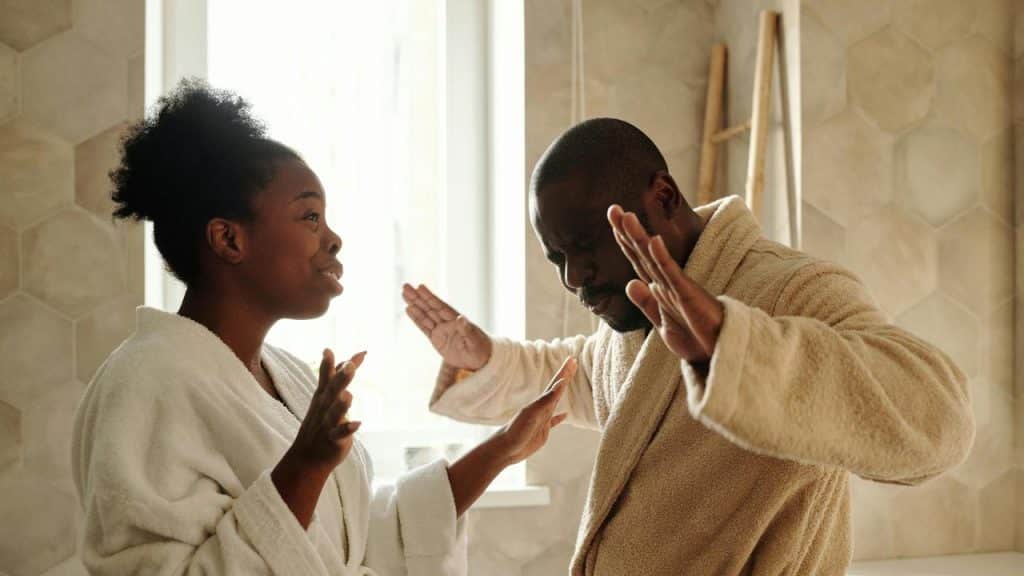 A man and woman in bathrobes arguing with raised hands in a bathroom.