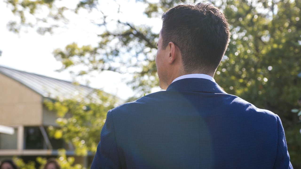 A view from behind a man in a blue suit looking toward trees and a building.