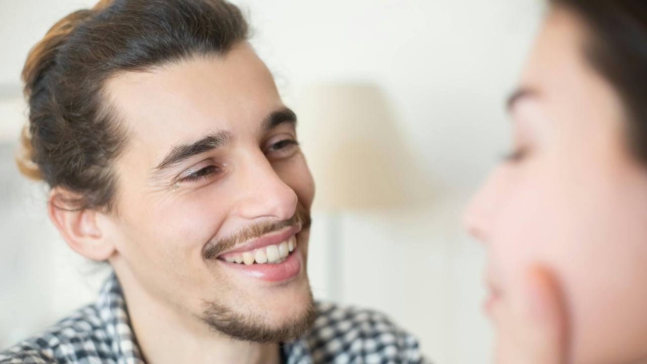 A close-up of a smiling man with a beard and bun looking at a person off-camera.