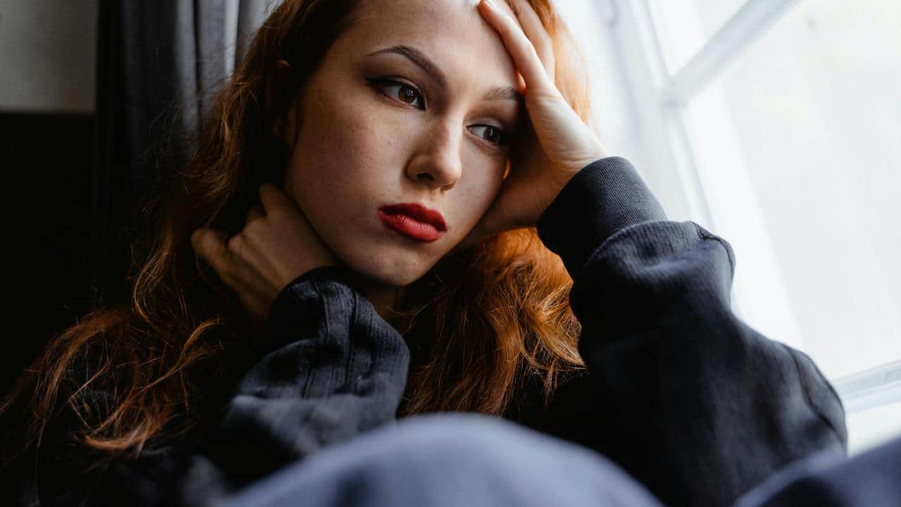 A woman holding her head, looking worried while sitting by a window.