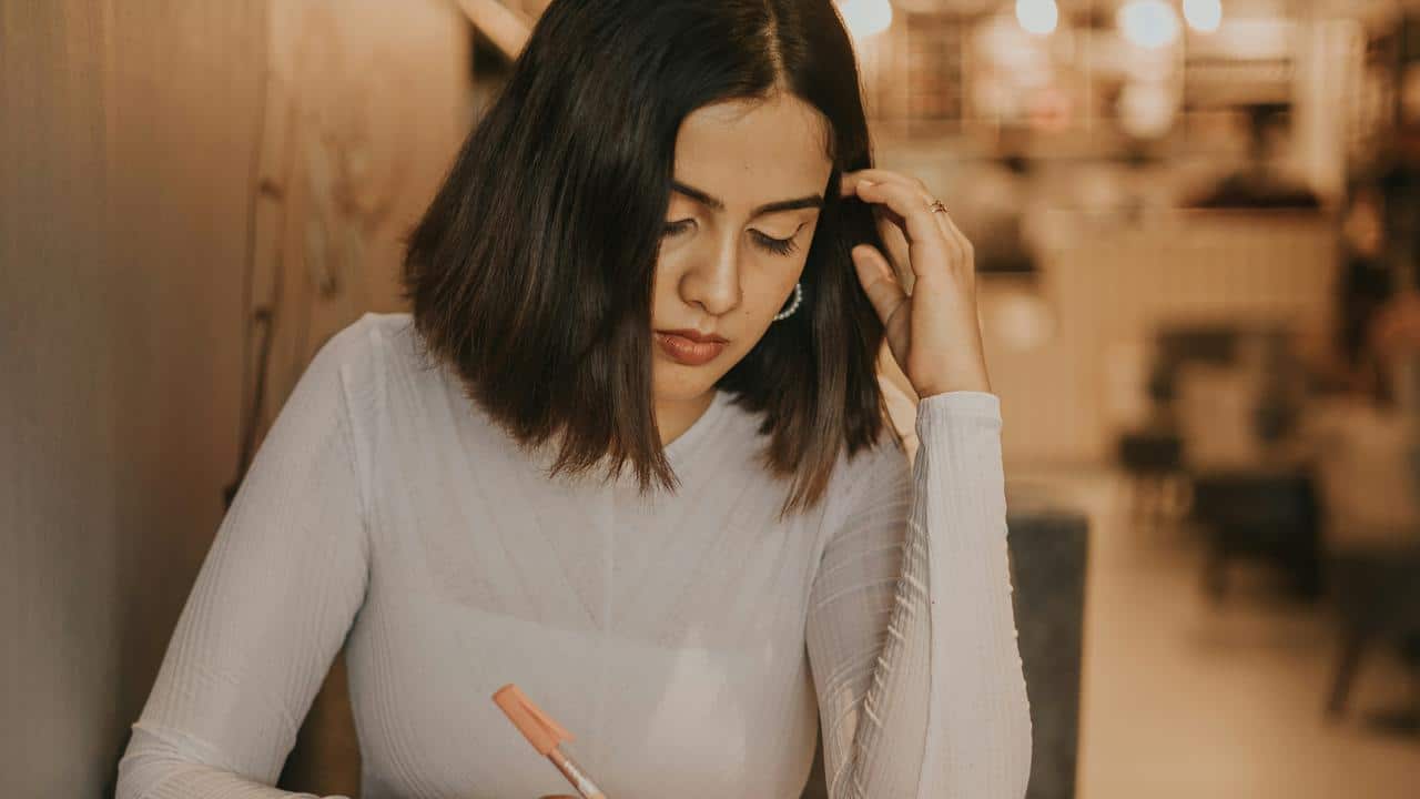 A woman writing thoughtfully at a table in a softly lit cafe.