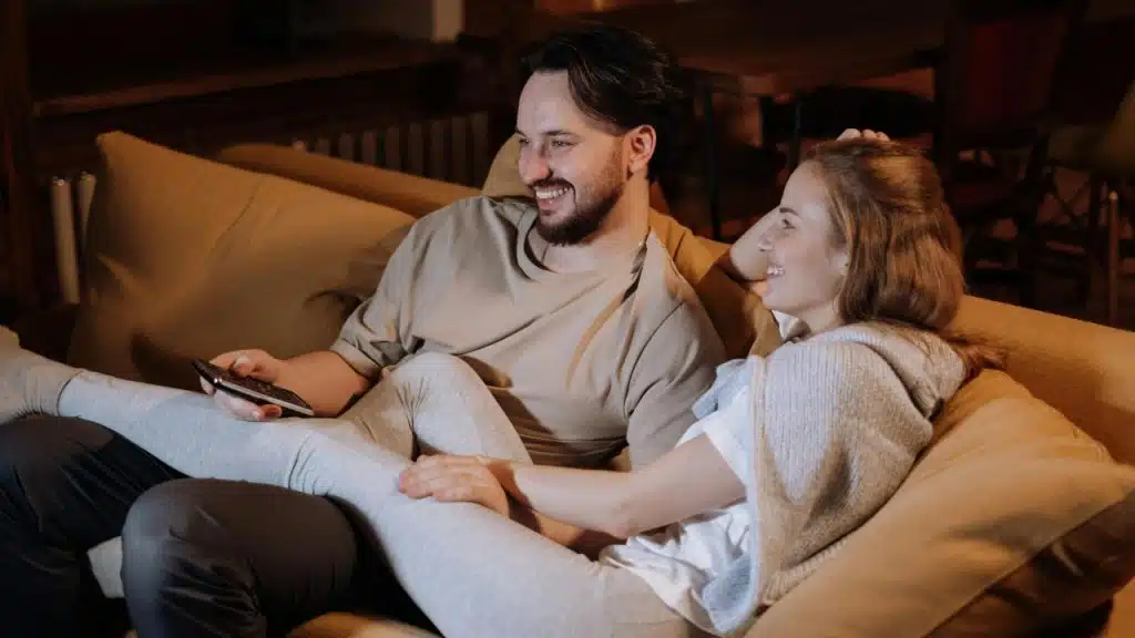 A man and woman sit on a tan sofa, smiling and holding a remote control.