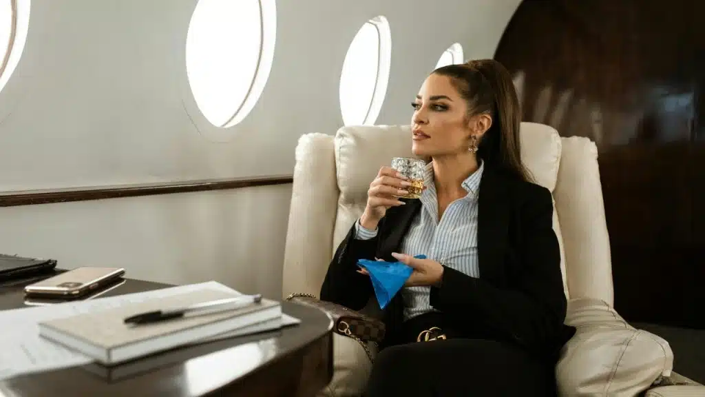 A woman in a black blazer sits in a leather plane seat holding a glass.