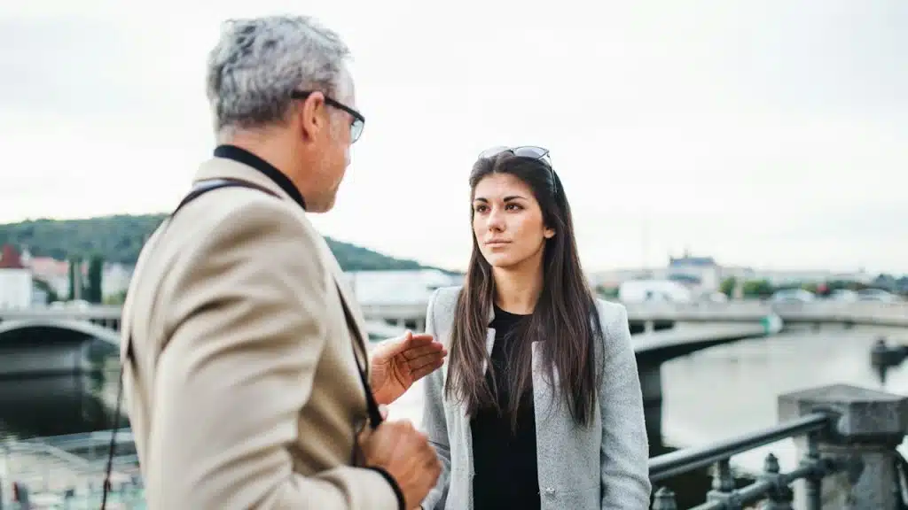 A mature man and a young woman are walking by a river in the city of Prague talking.