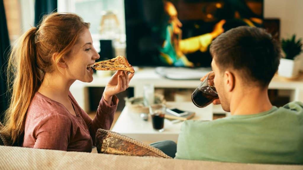 A couple relaxes together on a couch, enjoying pizza and drinks while watching TV.