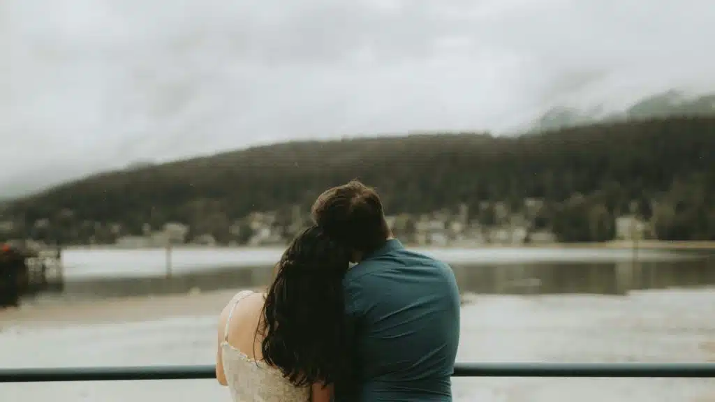A couple leans on each other while gazing at a calm lake surrounded by mountains.