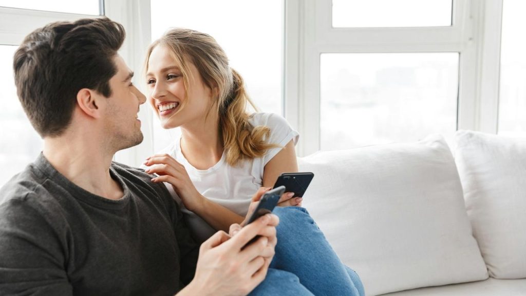 A smiling couple sitting on a couch with smartphones.