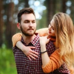 A bearded man carrying a woman on his back while they stand in some woods.