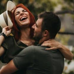 A man is holding up his wife, who’s holding a hat in one hand.