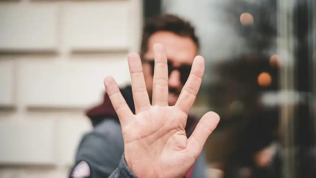 A close-up of a hand held up with five fingers spread, palm facing forward, with a blurred man in the background.