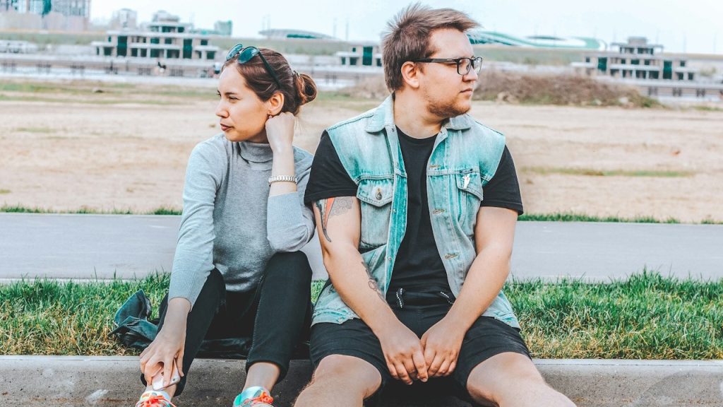 A woman looking at her right is sitting at the curb beside a man looking at his left.