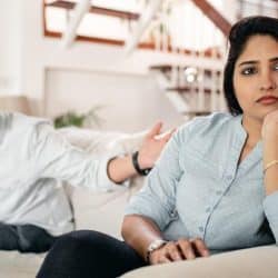A woman is sitting on a couch with a distant expression while a man in a turban sits behind her, gesturing toward her.