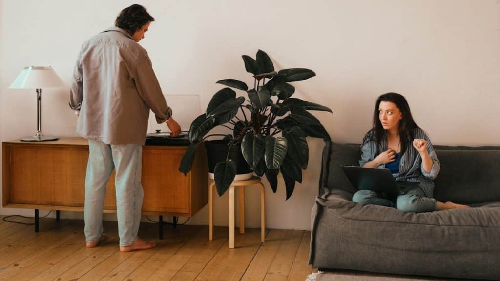 A woman is working on a laptop while her husband is standing near her.