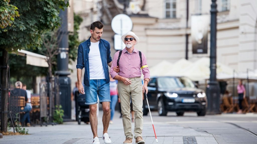 A young man and a blind senior with a white cane walking on pavement in the city.