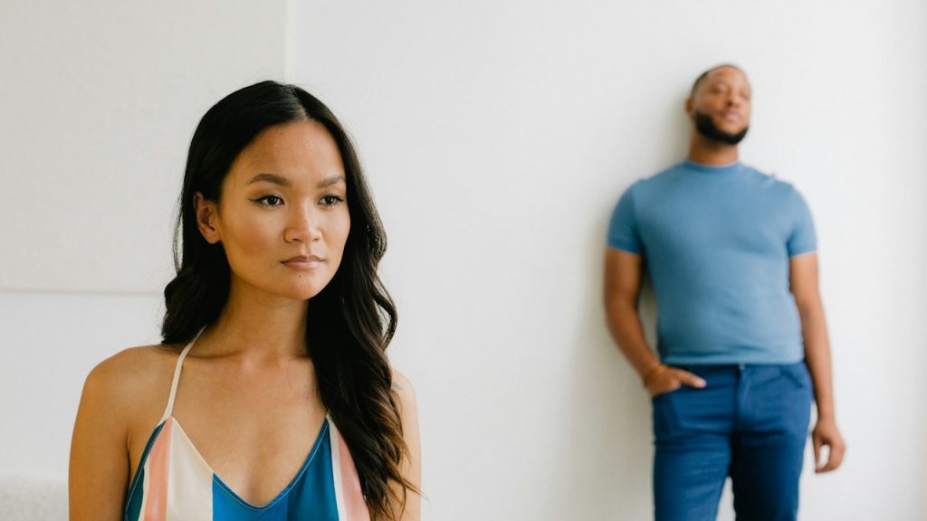 A thoughtful woman is standing, and her husband is leaning on the wall while standing behind her.
