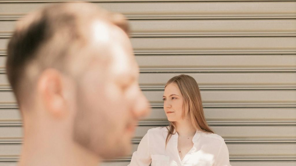A woman leaning a shutter of a garage, looking aside, while a man with blurred face is standing in front of her.
