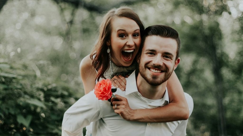 A woman holding a rose is riding on her husband’s back while they are walking through a forest.