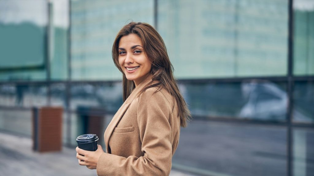 A woman holding a cup of coffee is posing for a picture outdoors.