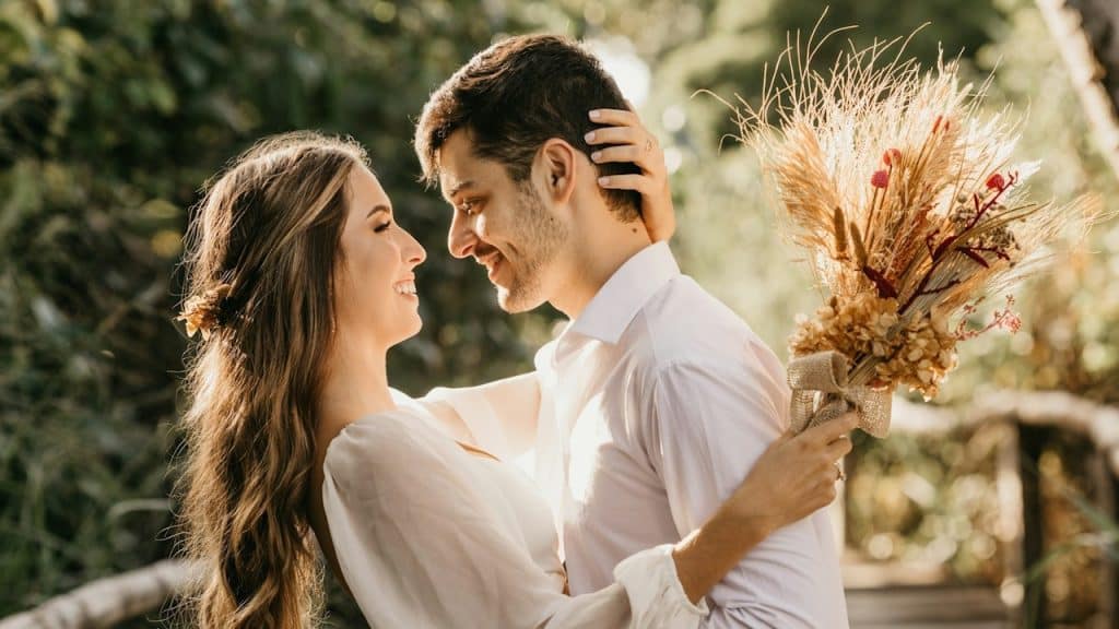 A woman in a white dress shirt holding a brown flower bouquet is hugging a man.