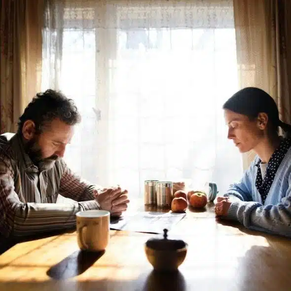 A man and woman are sitting silently across from each other at a sunlit wooden table, both looking down.