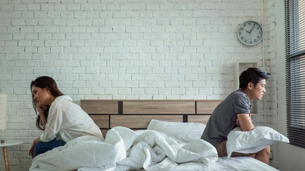 A couple sitting on separate ends of a bed facing away from each other