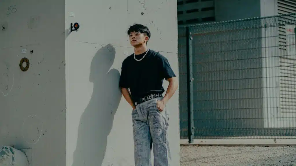 A man in a black crew-neck t-shirt and blue denim jeans standing beside a white wall.