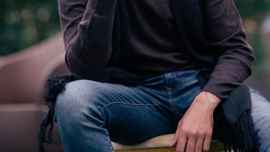 A man wearing a scarf and denim pants sitting on a chair.