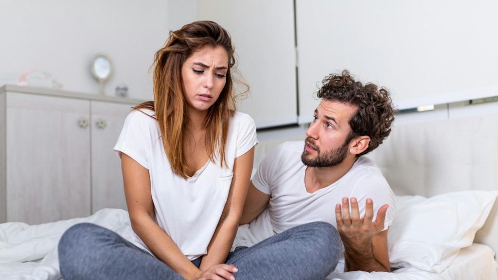 A man conversing with his upset wife while sitting on their bed.