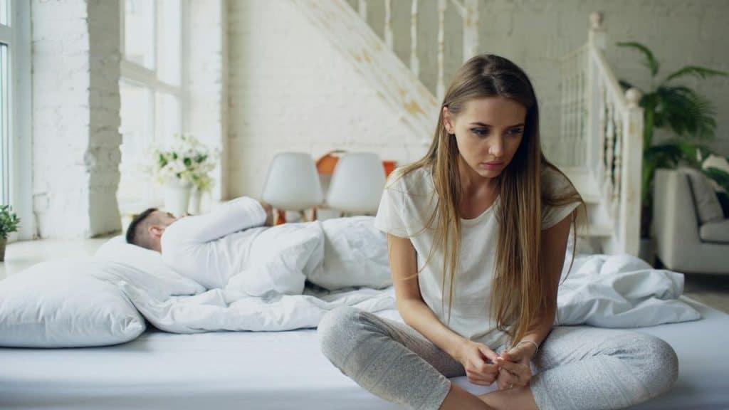 A young woman with a distressed expression sits cross-legged on a bed, while a man lies asleep under the covers in the background.
