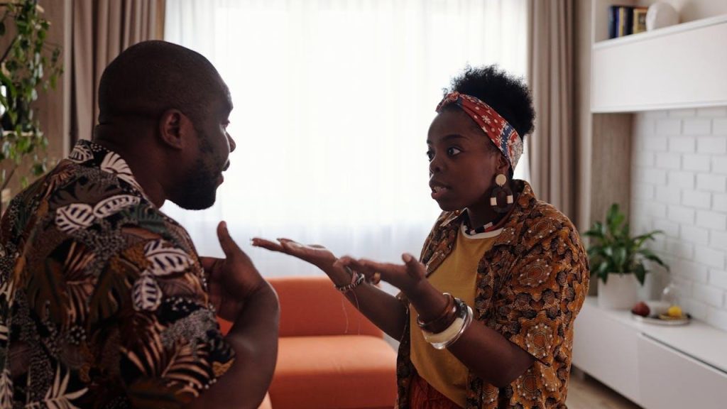 A man and a woman, both wearing patterned shirts, stand indoors facing each other and are gesturing during an apparently heated argument.