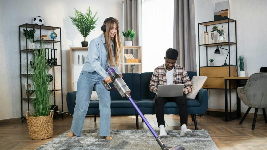 A woman wearing headphones is cleaning the carpet while her husband is working on a laptop.