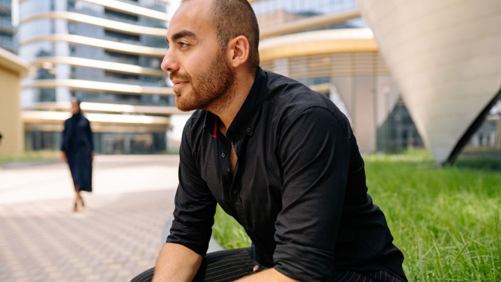 A man in a black dress shirt sitting on concrete near green grass.
