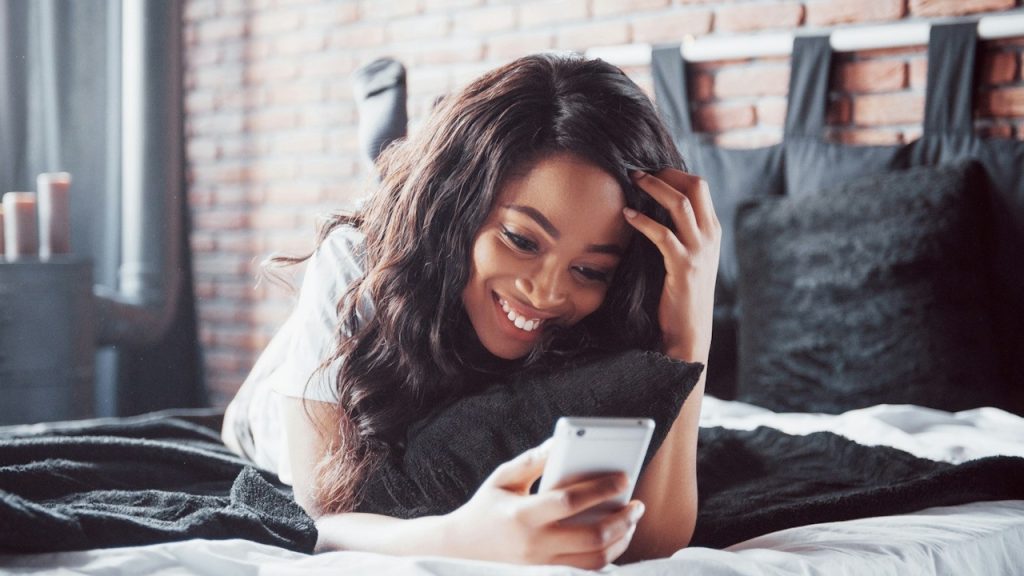 Woman lying on her stomach in bed, smiling at her phone.