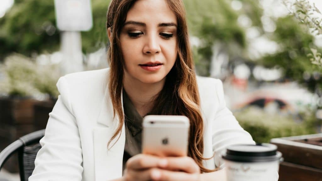 Woman in a white jacket using a mobile phone outdoors next to a coffee cup.