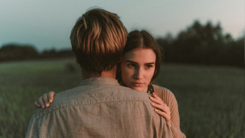 Woman looking seriously at the camera over a man's shoulder in an embrace outdoors.