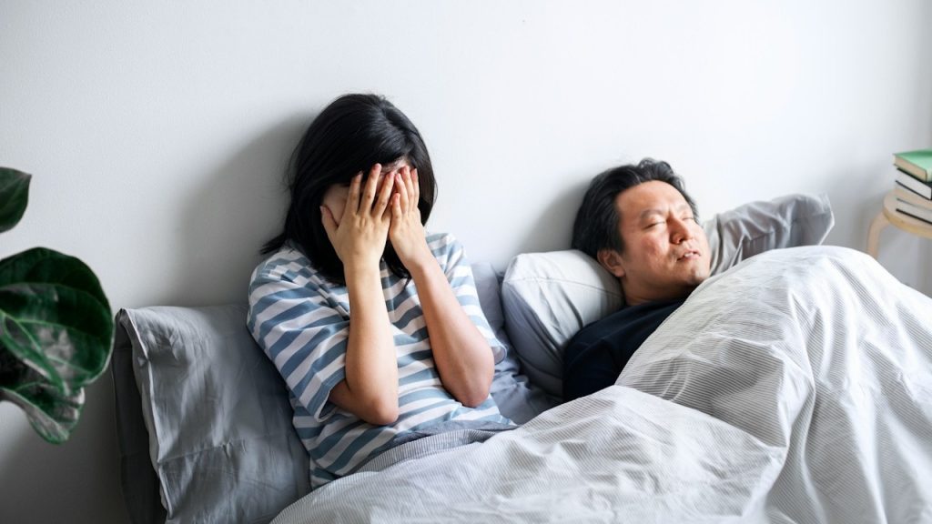 Woman sits up in bed covering her face with her hands, while a man sleeps next to her.