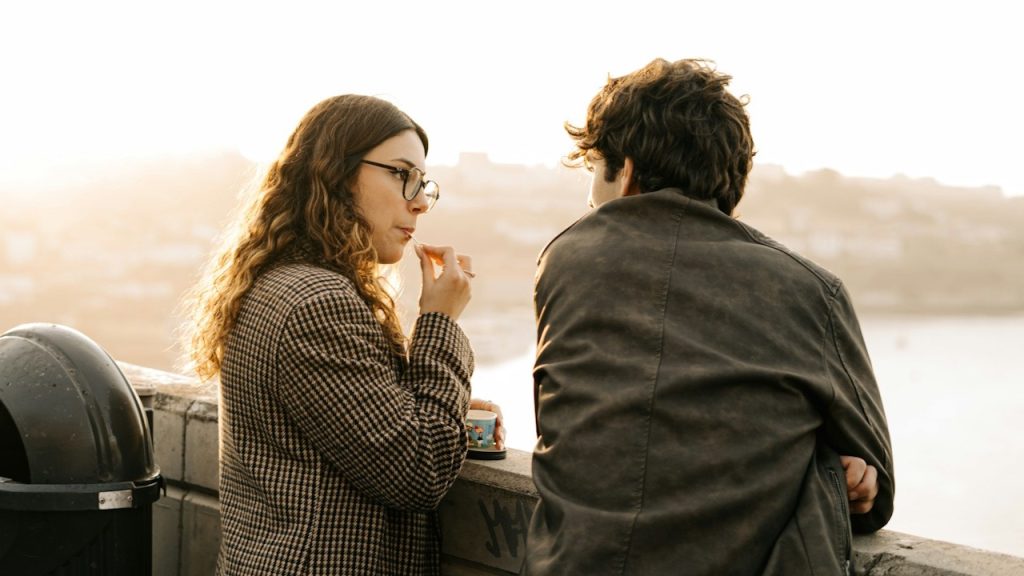 A woman in glasses and a checkered coat talks to a man while leaning on a railing at sunset.