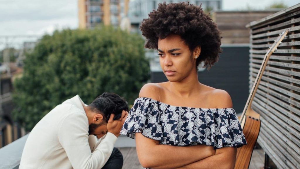 Woman with arms crossed kneels on a rooftop picnic blanket, looking upset while a man sits behind her with his head in his hands.