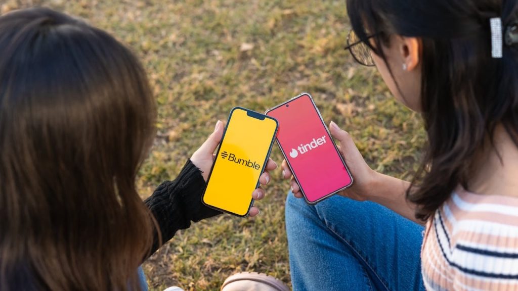Two women sit outdoors, each holding a smartphone displaying the logos for the dating apps Bumble and Tinder.