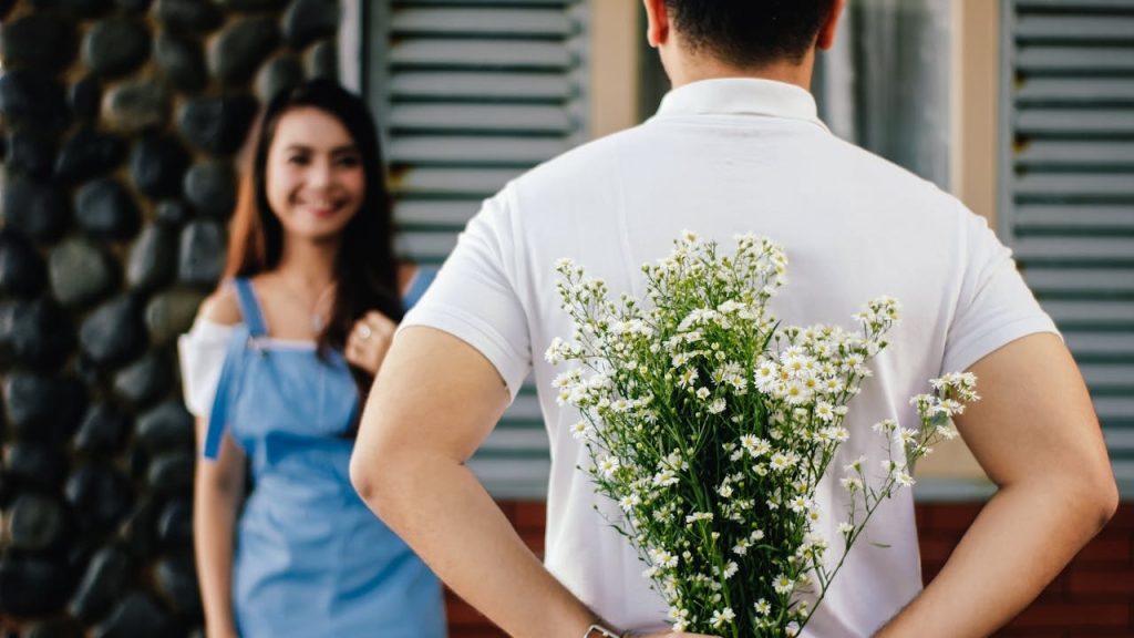 Man hides a bouquet of white flowers behind his back while a smiling woman in blue overalls stands out of focus behind him.