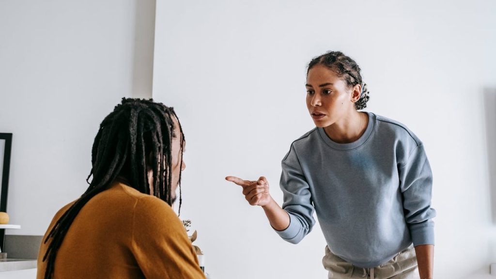 Woman with braided hair points a finger assertively at a man with dreadlocks sitting at a table.