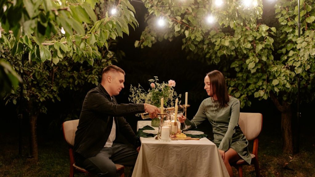 A couple shares a romantic dinner at night under string lights in a garden setting, toasting with glasses.