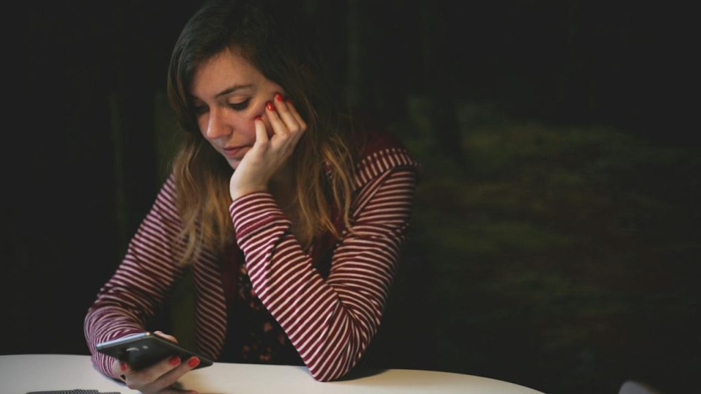 A young woman with her chin resting on her hand looking sadly at her smartphone