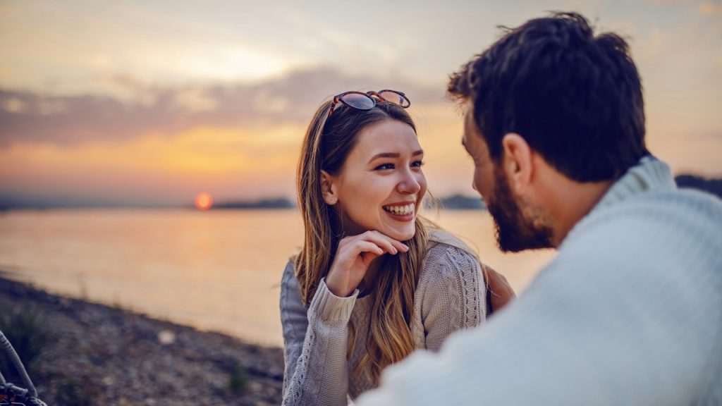A cute, smiling, Caucasian brunette is sitting on the coast near the river with her loving boyfriend and flirting in the background.