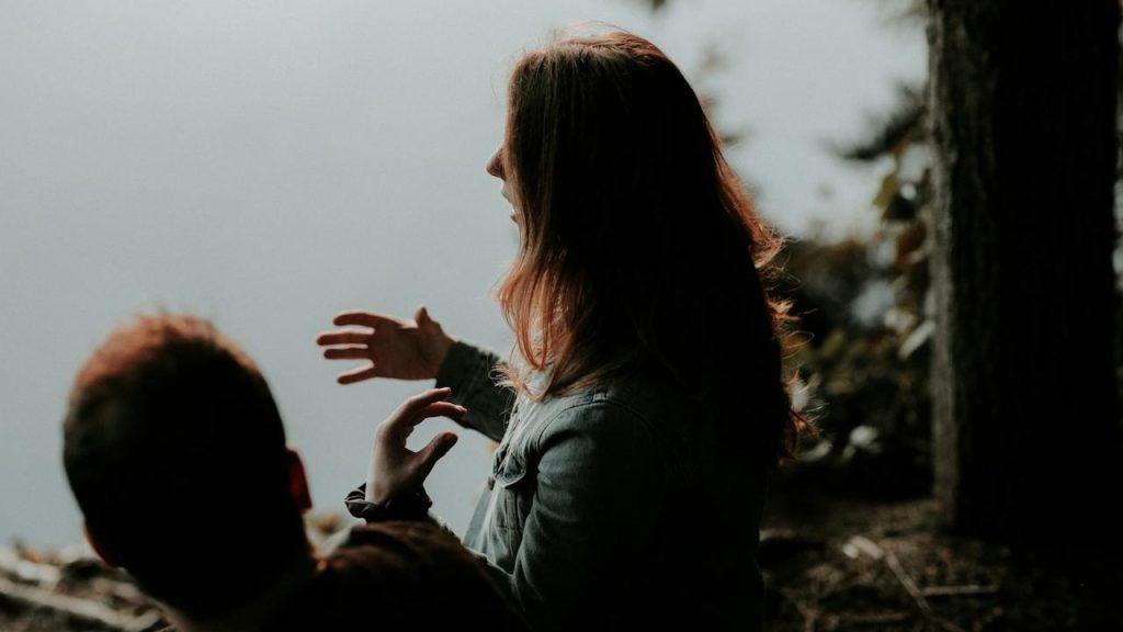 Woman gesturing with her hands while talking to a man outdoors by a foggy lake.