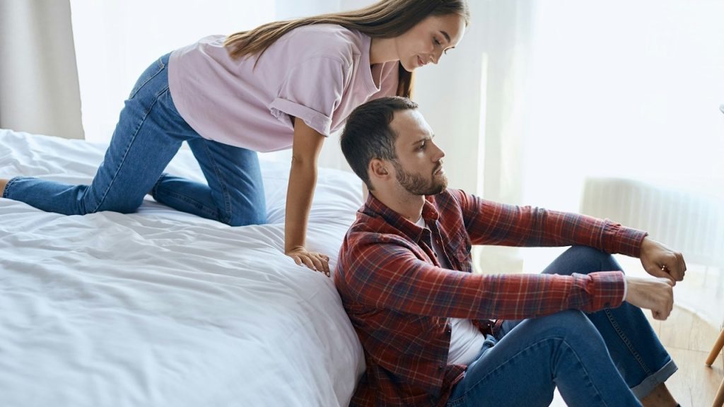 Man dressed in casual outfit sits on light coloured floor listens to news attentively pretty playful woman creeping up from behind stands on bed.