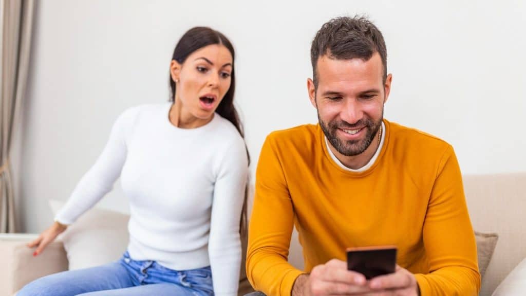 A man smiling as he uses his phone and his wife looks on in shock while they sit on a couch.