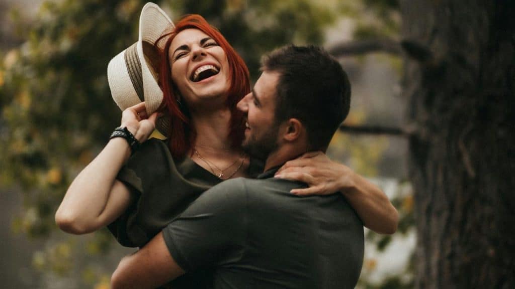 A man is holding up his wife, who’s holding a hat in one hand.